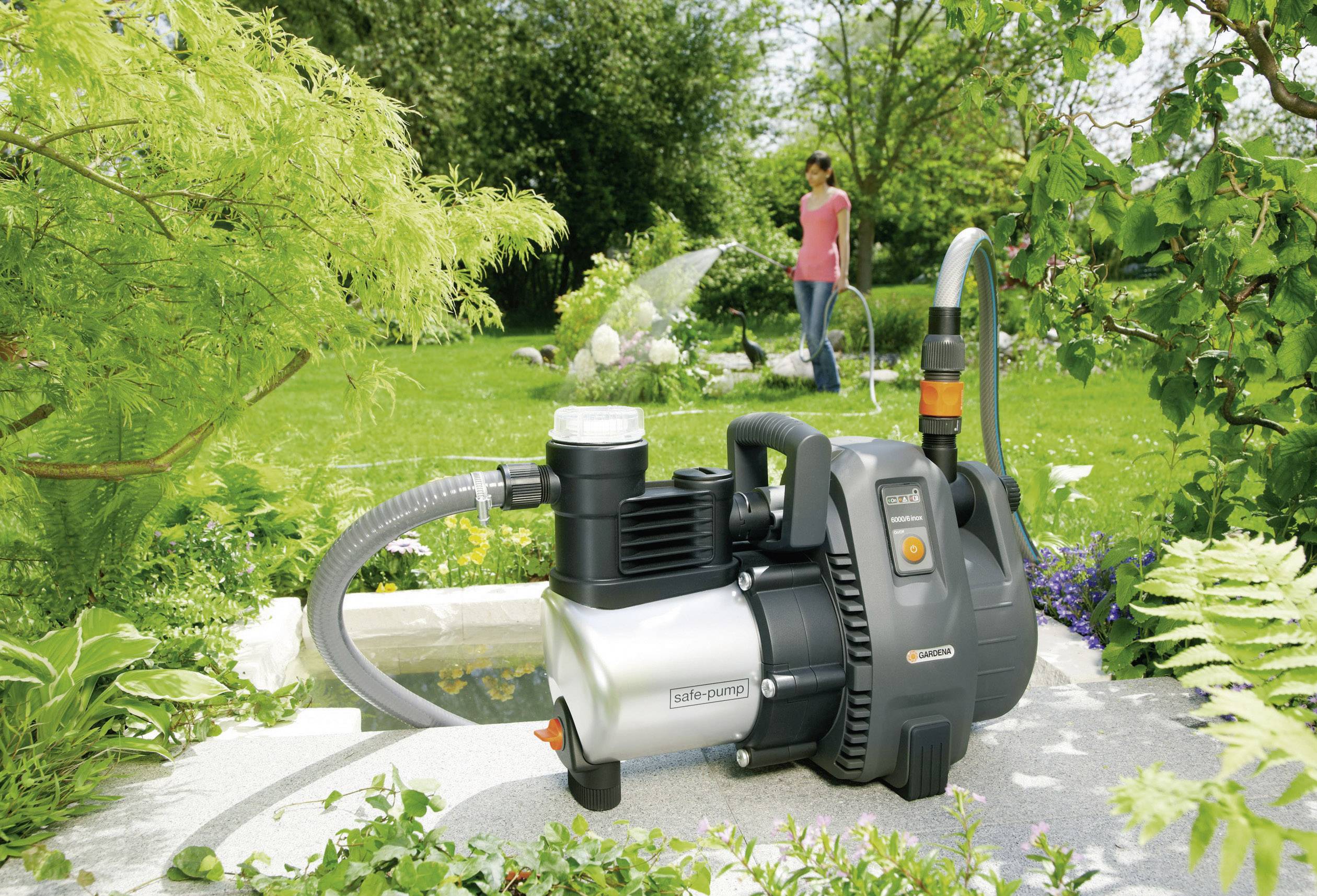 'Garden pump on stone, with green plants beside it. In the background, a person is watering the lawn with a garden hose.'