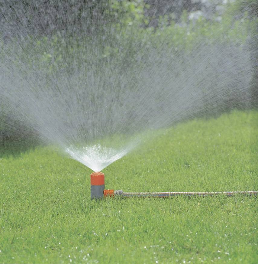 A lawn is being watered by a sprinkler. Water jets are distributing evenly across the green grass in the garden.