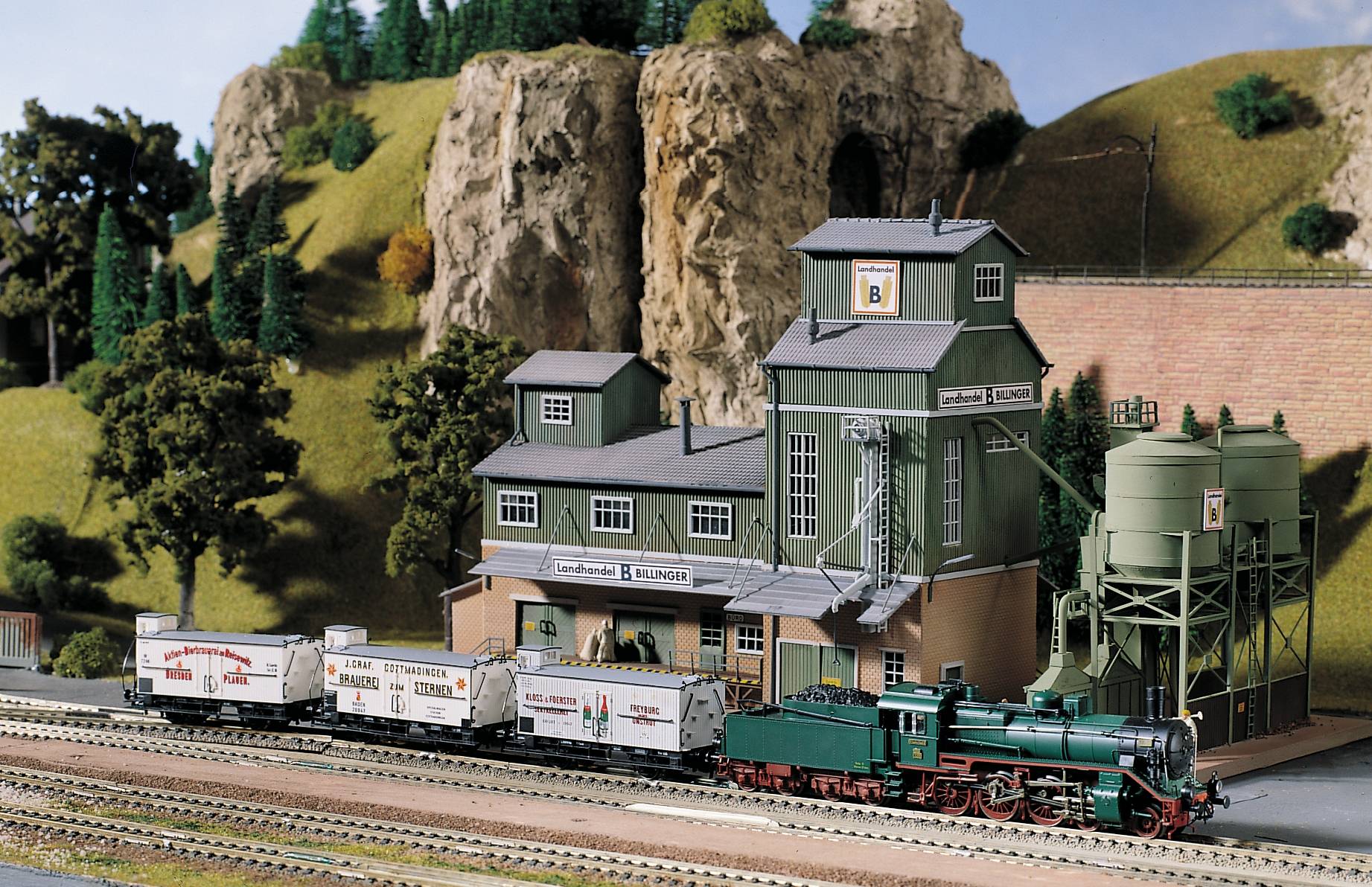 A model railway landscape with a steam locomotive on tracks, surrounded by hills and a building in the background.