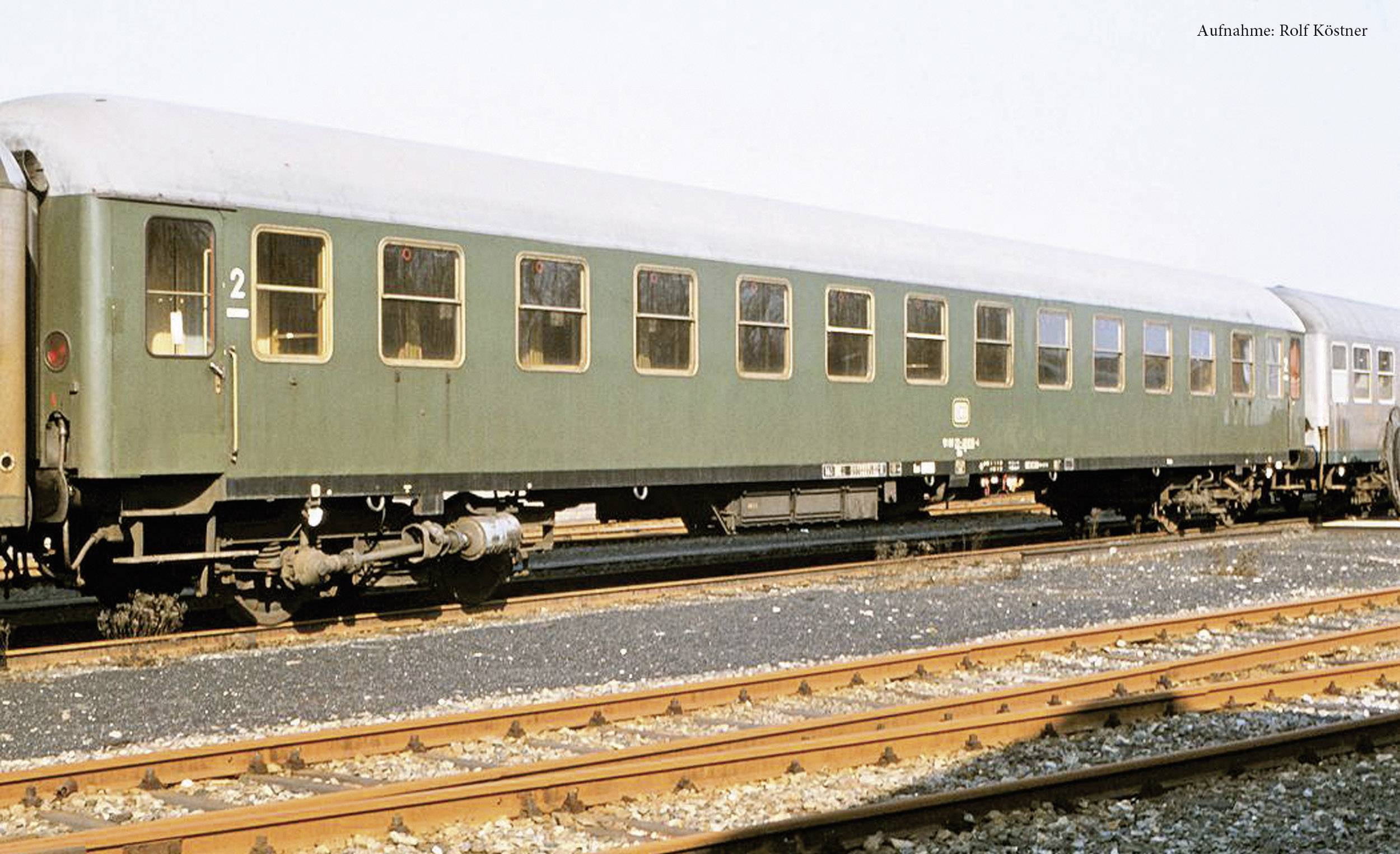 A green, old second-class railway carriage stands on a railway track on a sunny day.