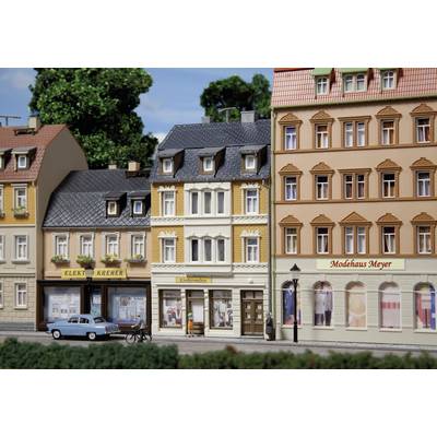 The illustration depicts a row of model buildings at the scale of a miniature village, including an electrical store and a fashion house.