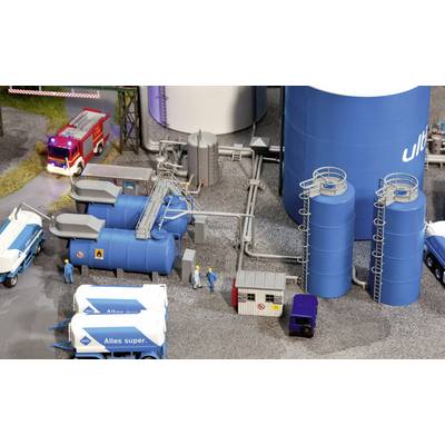 A miniature installation depicts an industrial scene with blue tanks, a red fire engine, and workers wearing blue overalls.