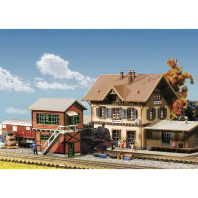 A miniature model railway station with two buildings, a train and figures. The sky in the background is blue with white clouds.