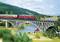 A red locomotive pulls a passenger train across a steel bridge, with a river flowing beneath. Cows are visible in the foreground.