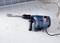 A blue hammer drill lies on a white tiled floor. Some tiles are damaged, suggesting construction work is in progress.