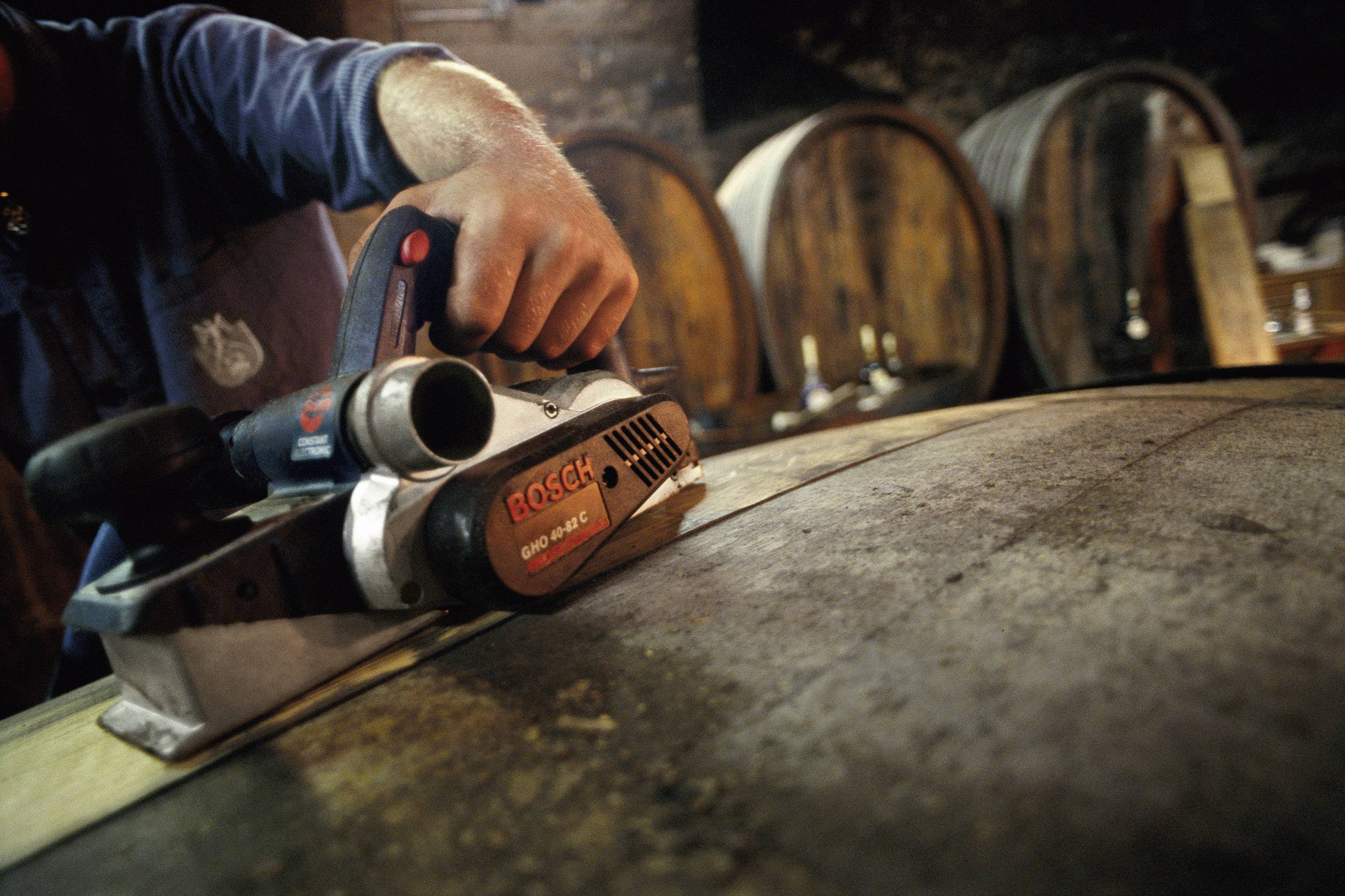 A craftsman smooths the surface of a wooden barrel with an electric tool in a workshop with large barrels in the background.