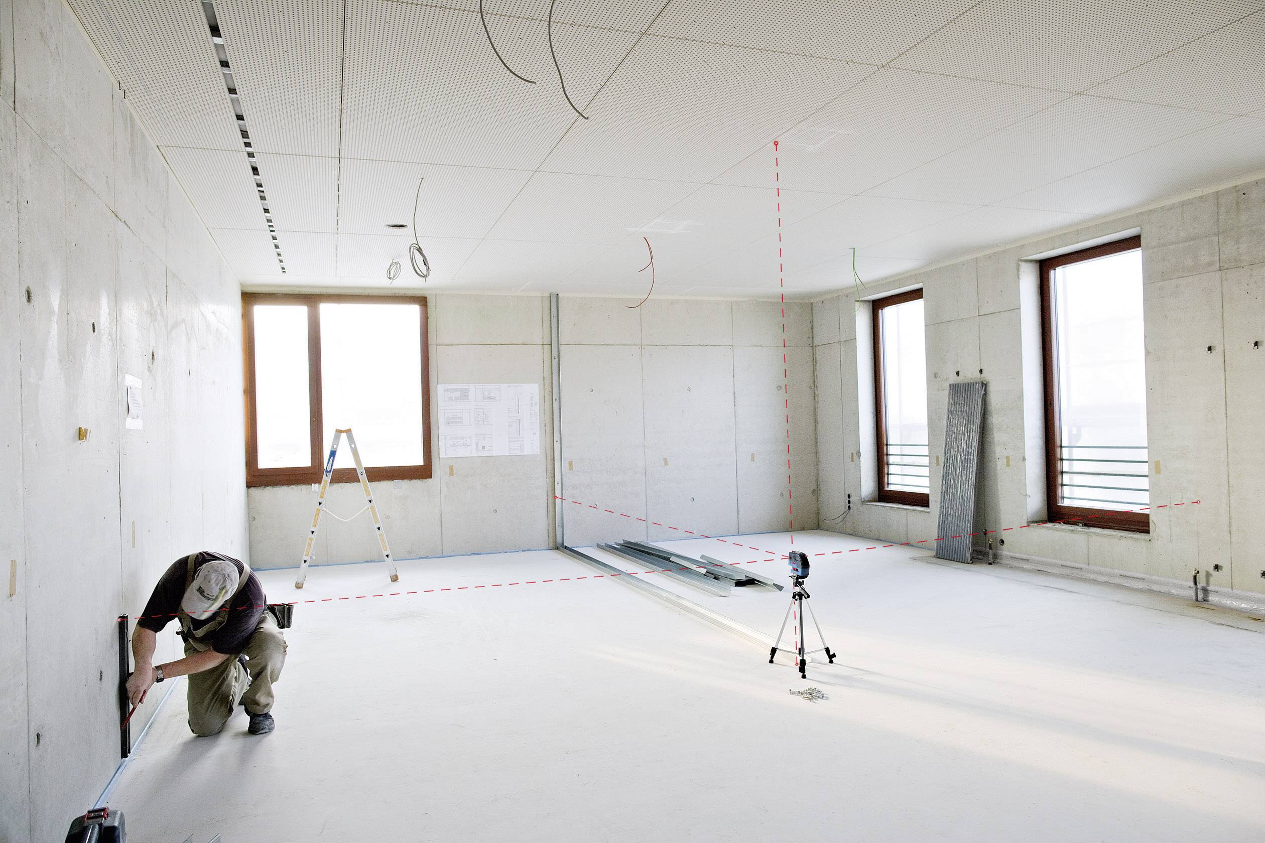 A man is measuring the distance in an empty room with concrete walls and ceiling. A ladder and construction plans are visible in the background.