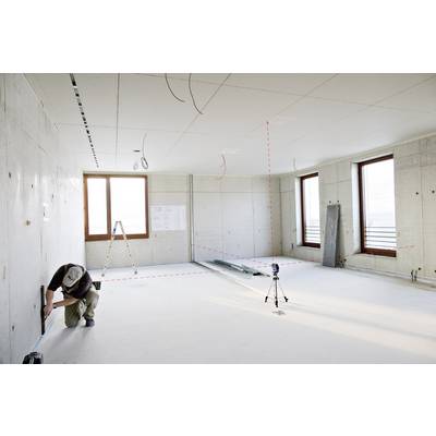 A man is measuring the distance in an empty room with concrete walls and ceiling. A ladder and construction plans are visible in the background.