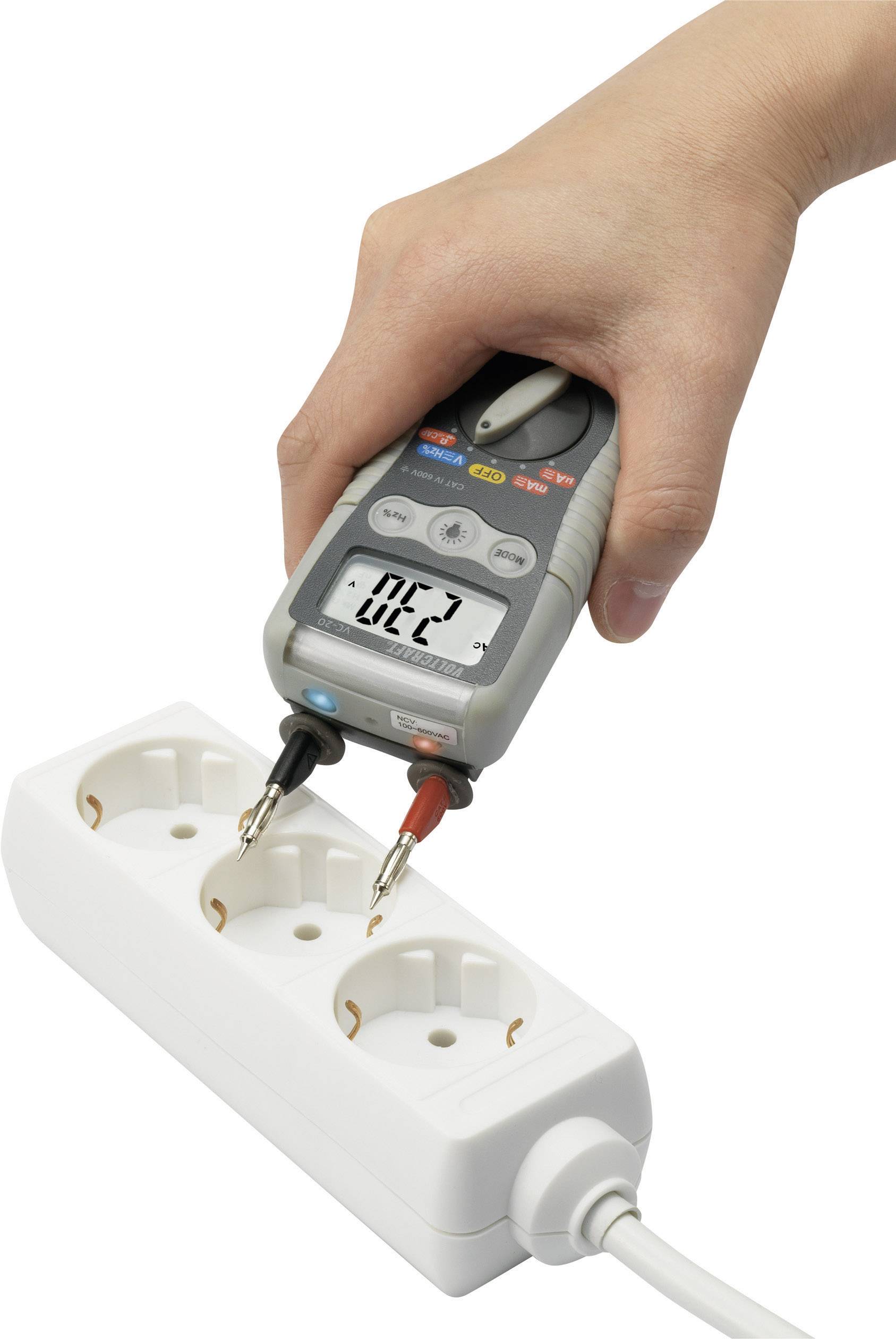 A hand is holding a voltage tester plugged into a white power strip, displaying a reading on the screen.