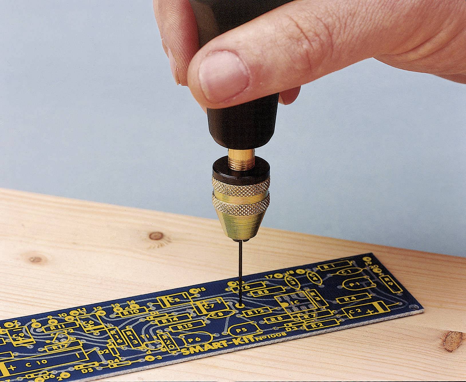 A hand holds a small drill and is drilling a hole into a blue circuit board lying on a wooden board.