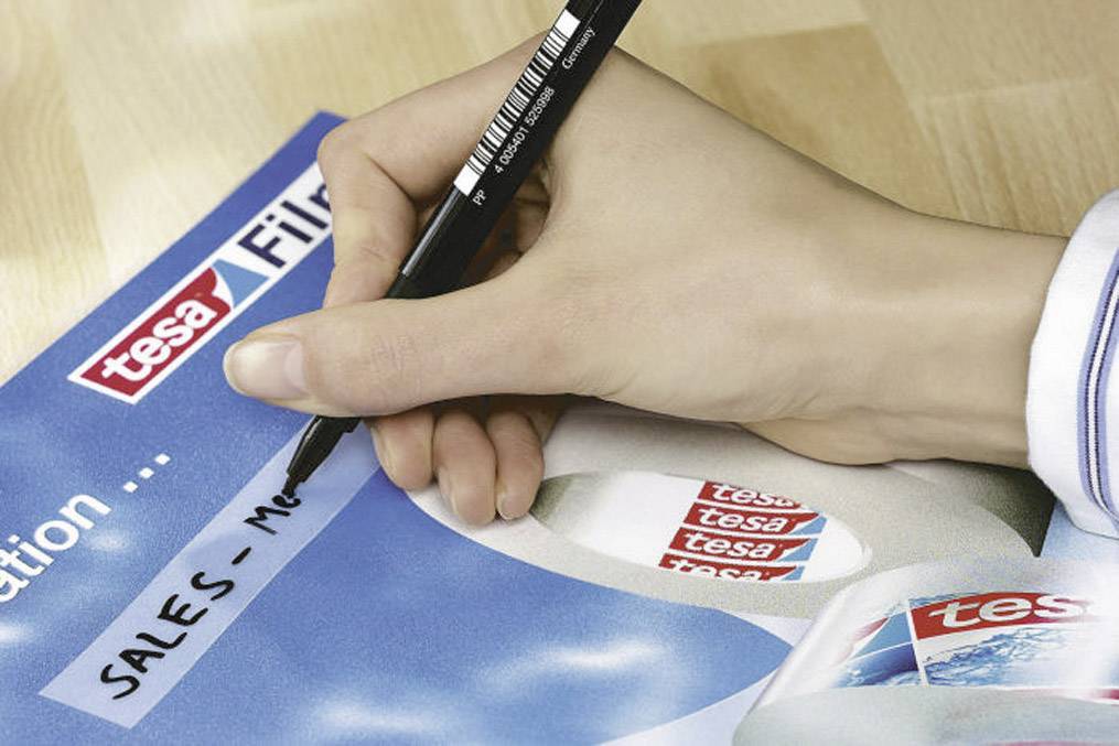 A hand is writing with a black pen on paper with a 'tesa' logo and the inscription 'SALES - MEETING'.