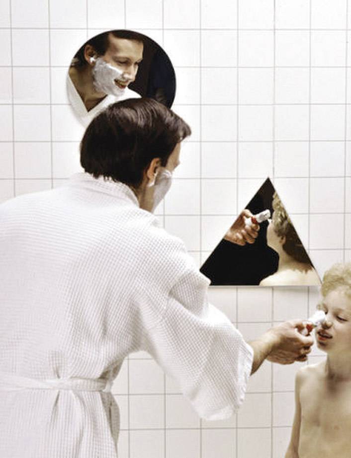 A man in a dressing gown is shaving in front of a mirror. In another mirror reflection, he observes a boy who is also being shaved.