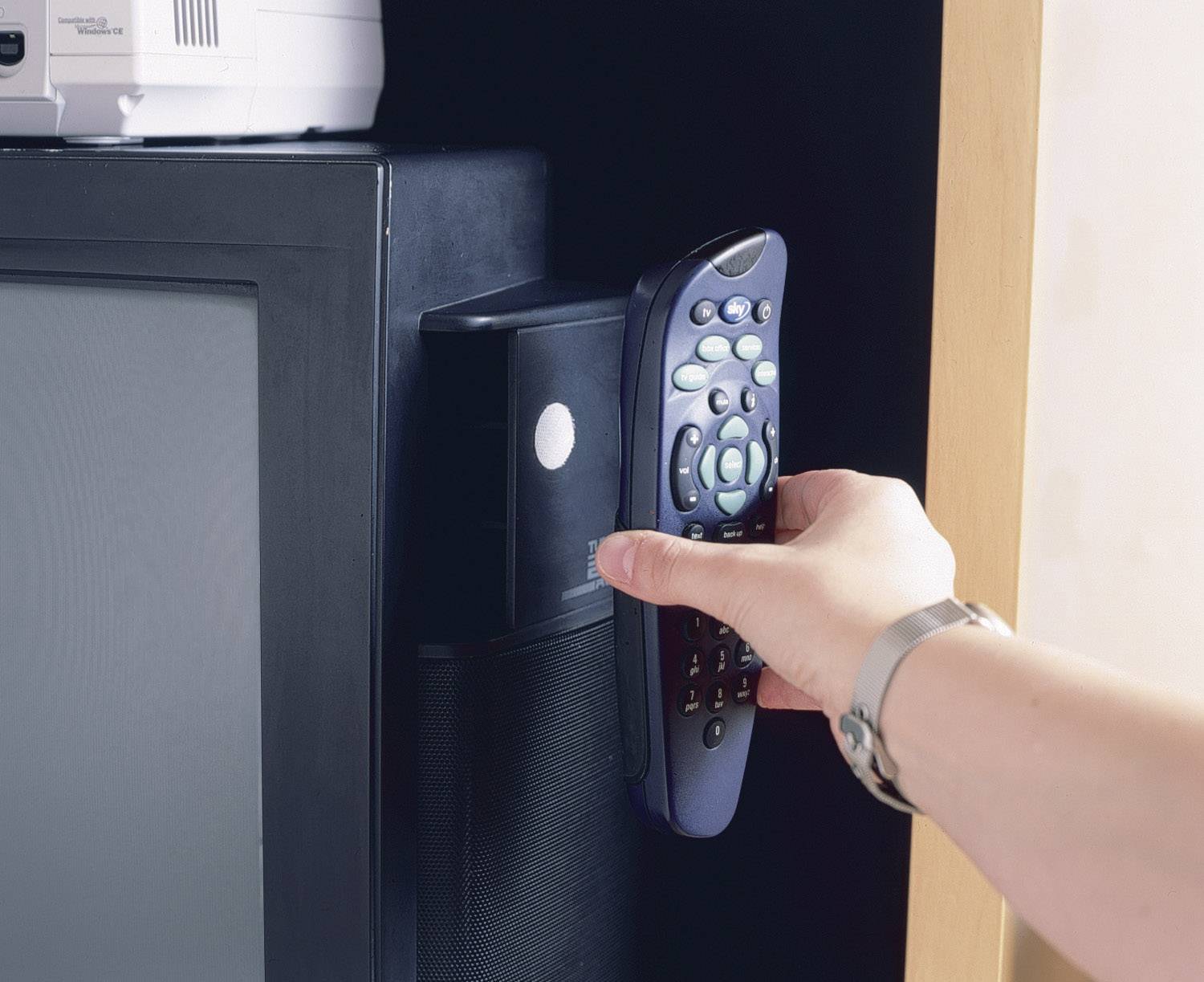 A hand is pointing a remote control at a black speaker box next to a television. The scene suggests the control of audio or video devices.