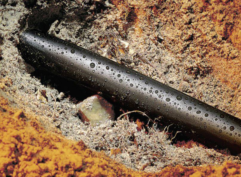 A black pipe with water droplets, embedded in an earth trench of brown and yellow sand.