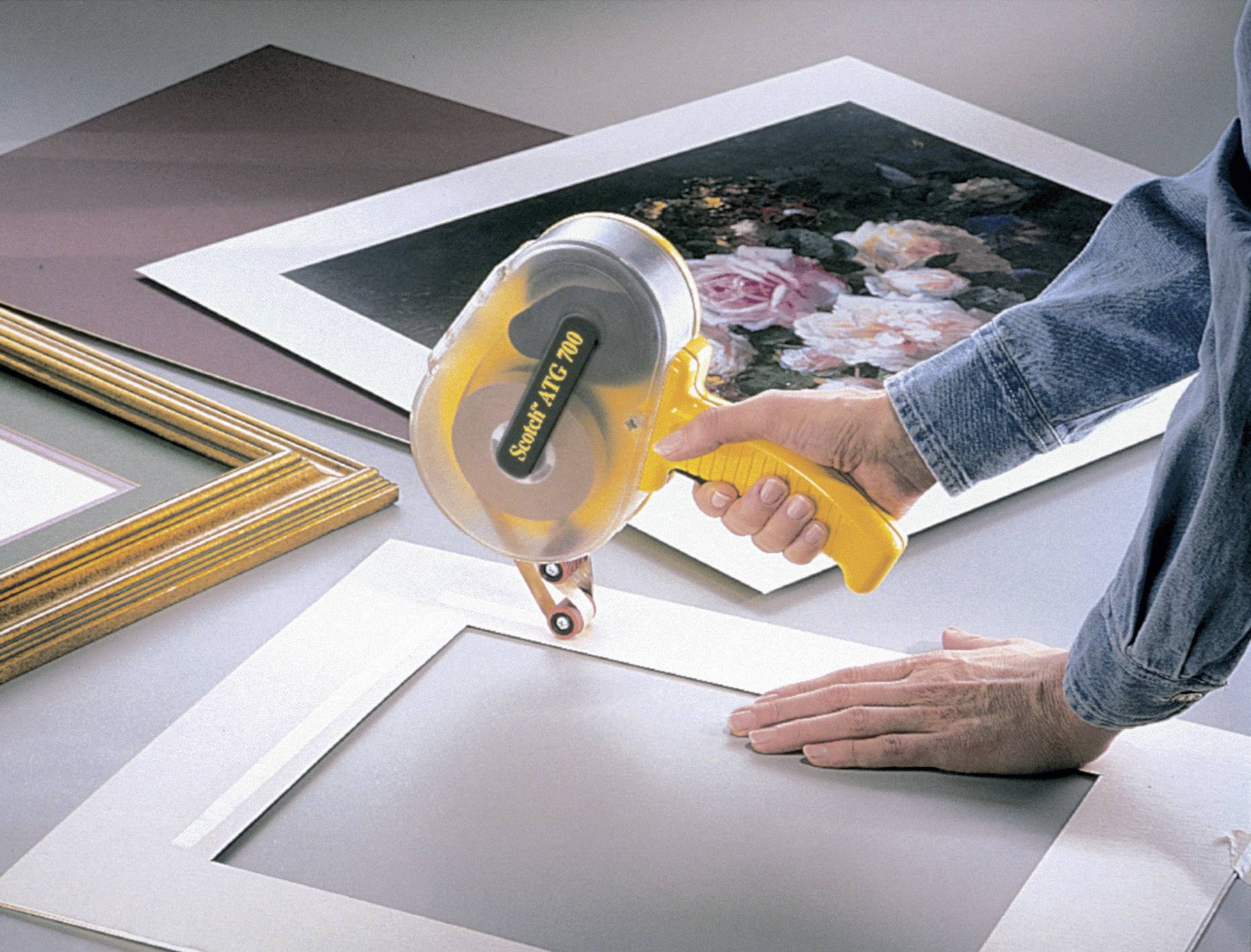 A person is using a yellow glue gun to attach a mount to a floral picture. Another frame lies beside it.
