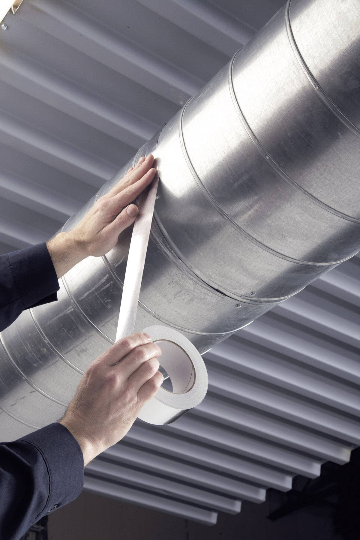 A person is applying silver adhesive tape to a large metal pipe in an industrial setting, in order to seal or secure it.