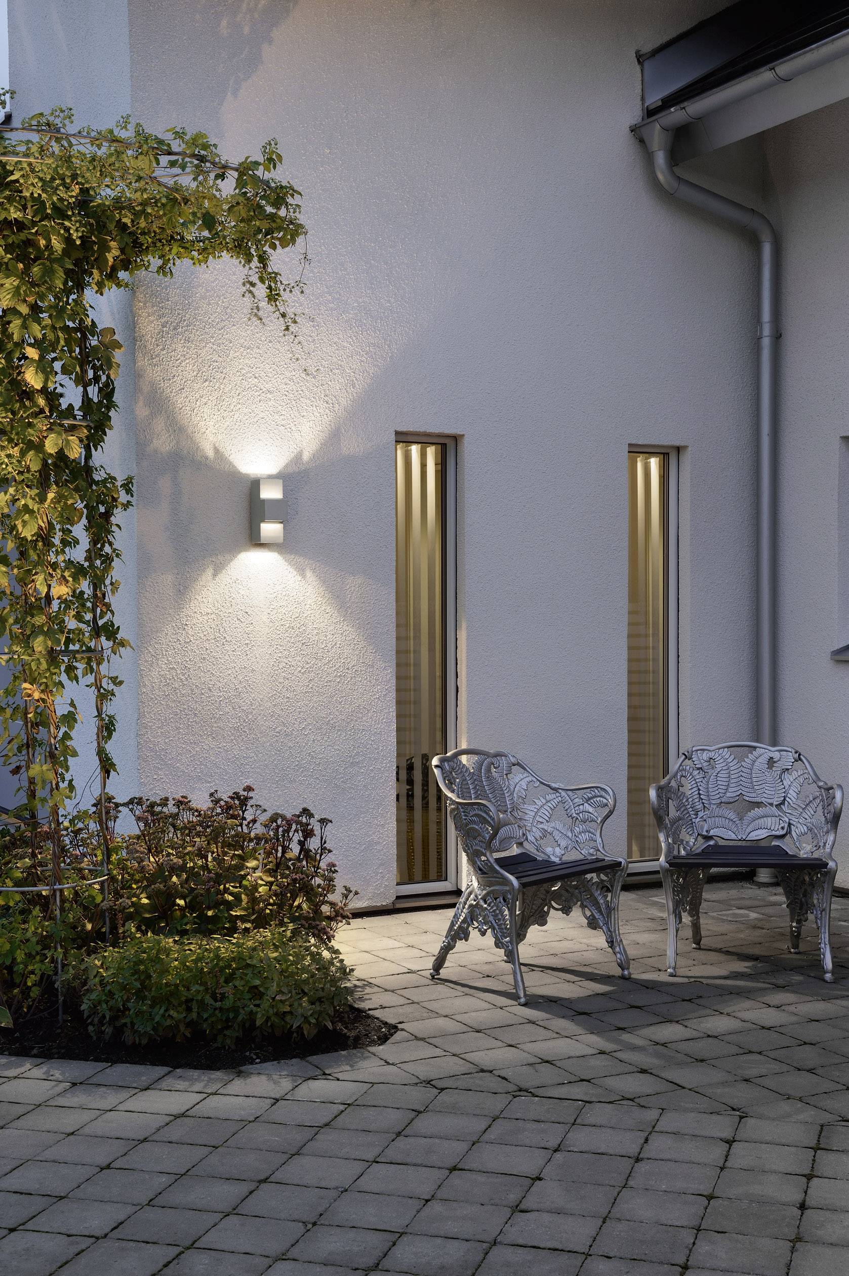 A lit outdoor area with two empty metal chairs on a paved floor. Climbing plants are growing on the wall.
