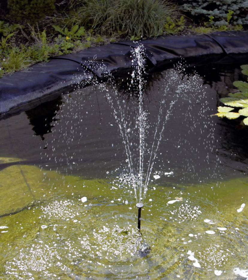 A small, bubbling fountain in a pond, surrounded by plants. Water droplets splash into the air, creating bright patterns.