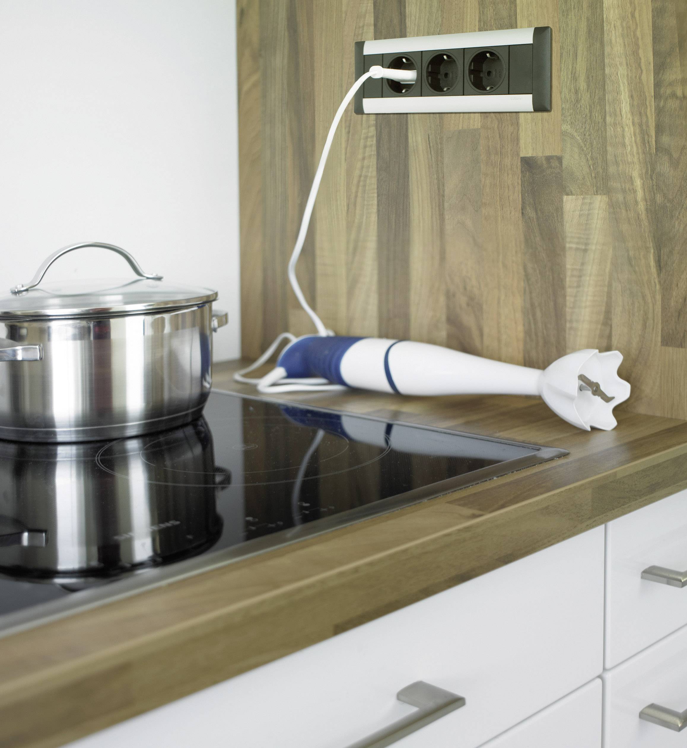 A saucepan on a hob next to a hand blender. A blender is plugged in, but not in use. The background shows a wooden worktop.