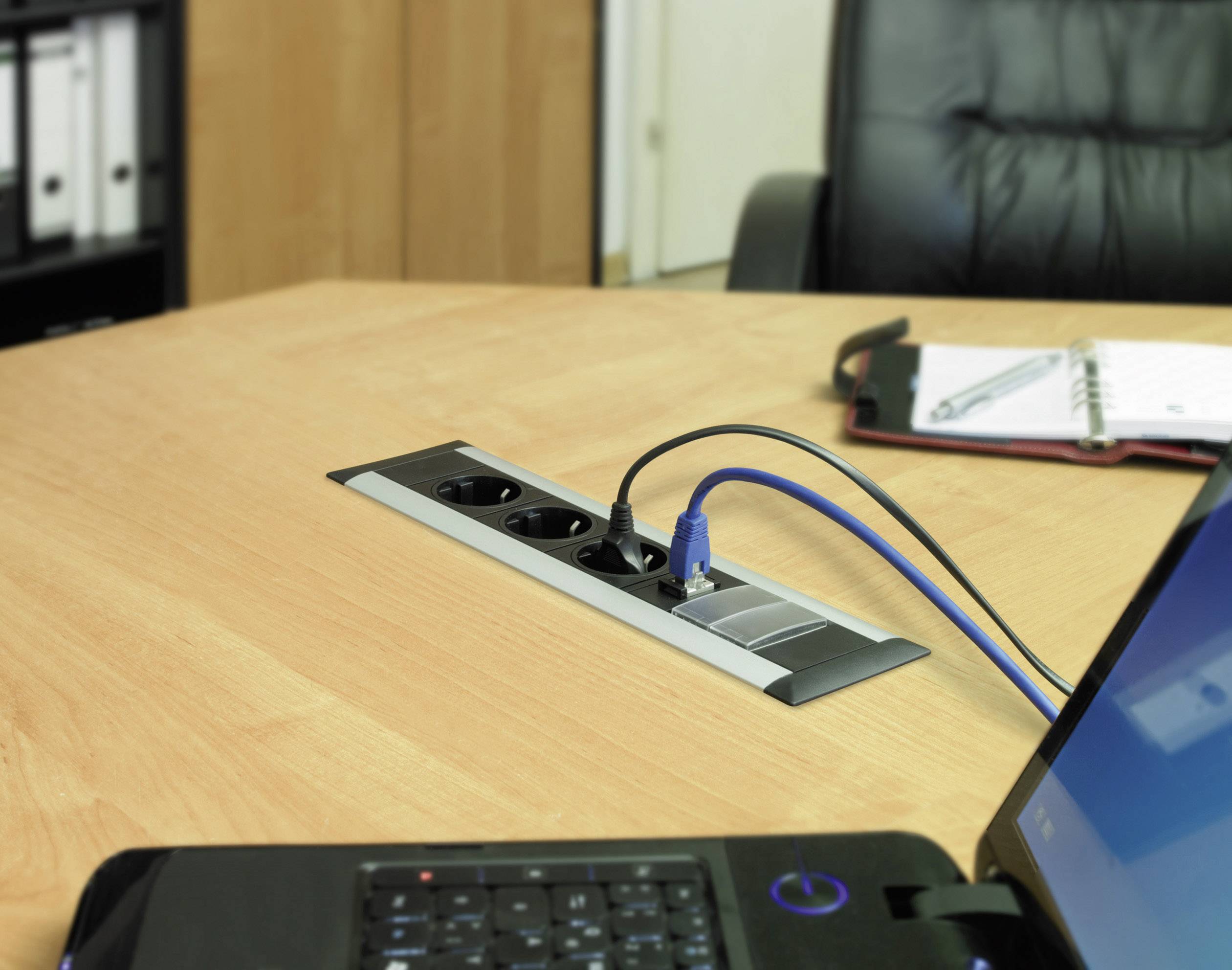 Laptop on a table, plugged into power sockets. A notebook lies beside it. Office environment in the background.