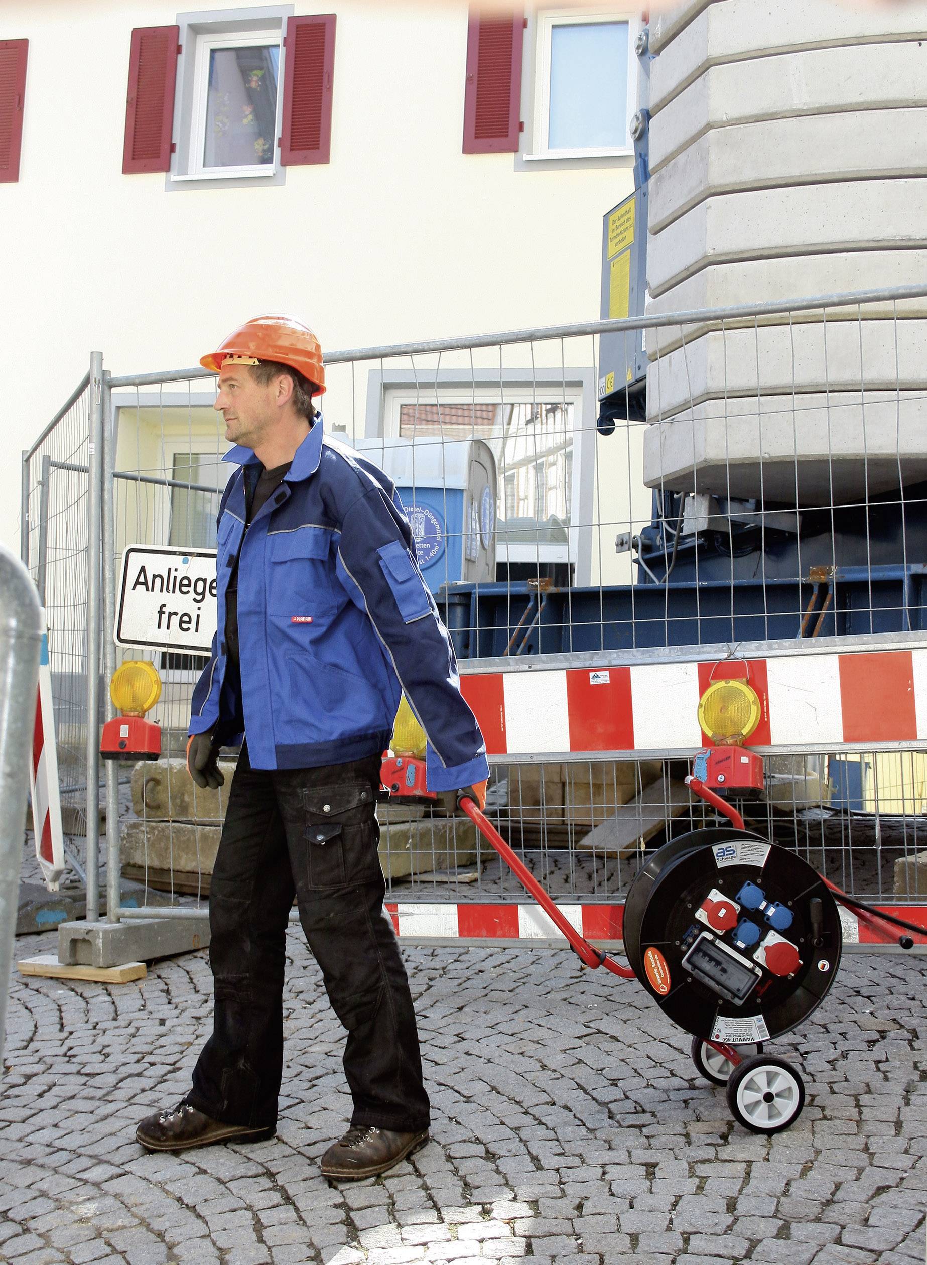 A construction worker in protective clothing is moving a large cable drum on a paved surface in front of a construction site with barriers.