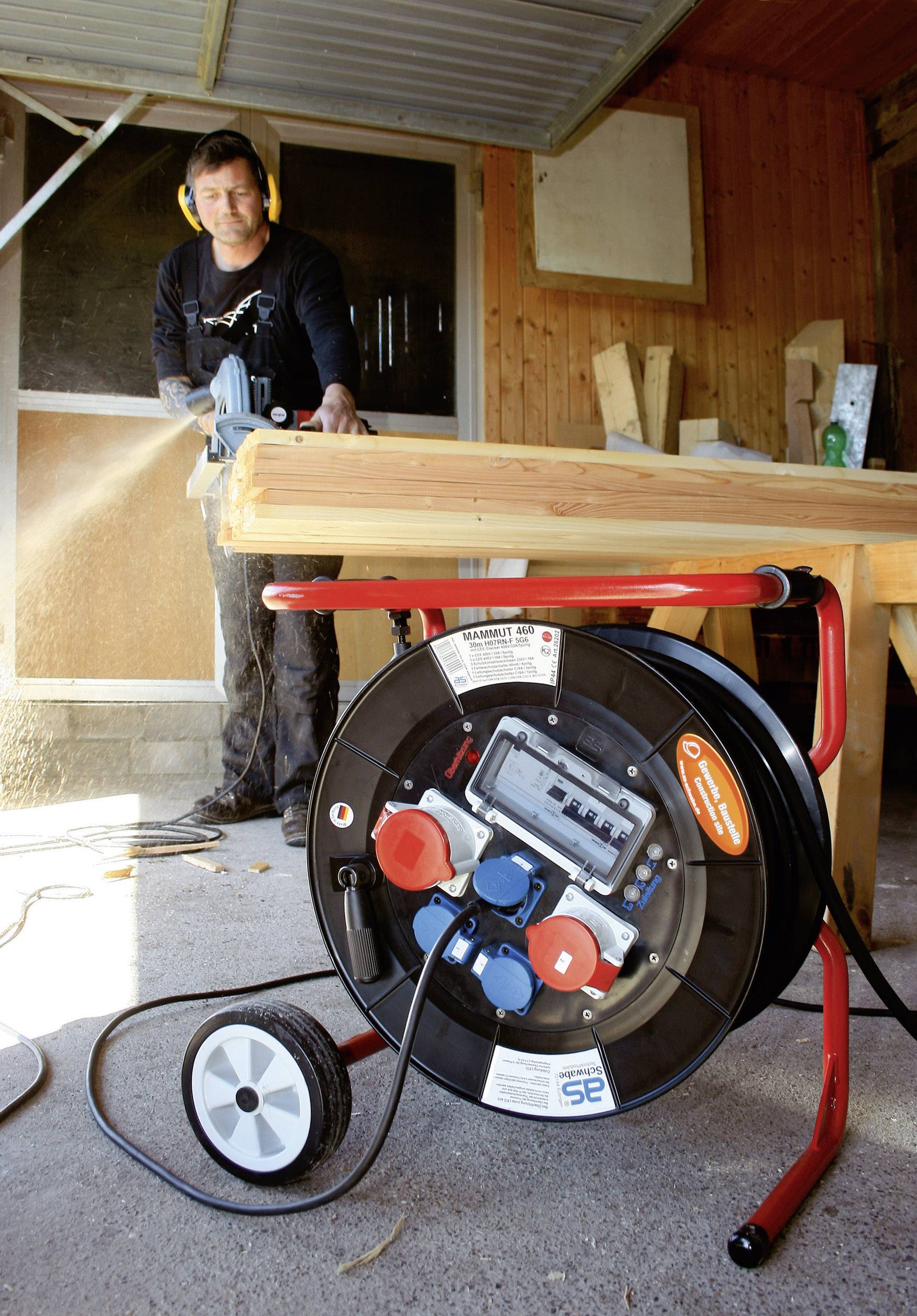 A man is wearing ear defenders and using a circular saw to cut a wooden board on a workbench. A cable reel is positioned in the foreground.