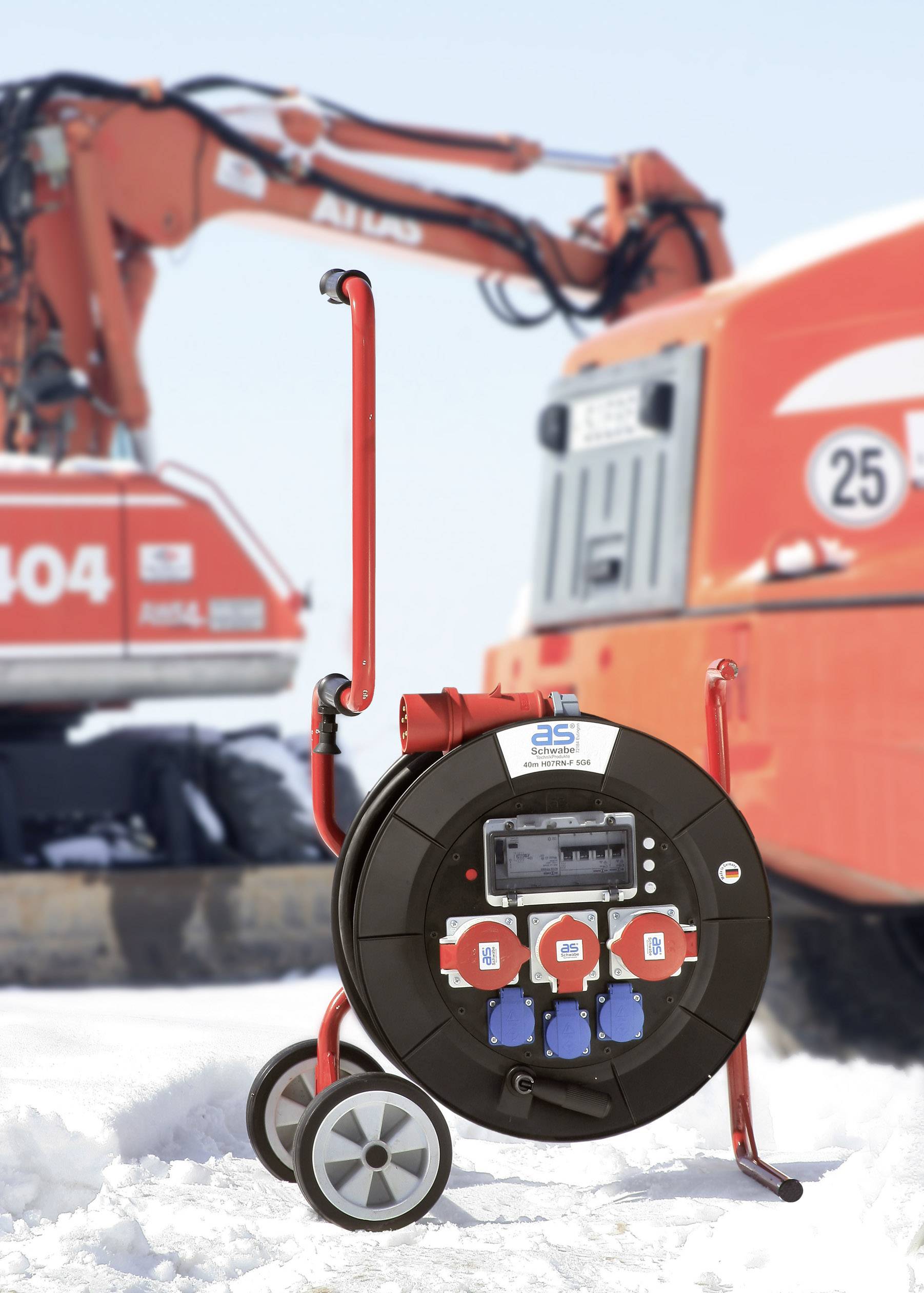 A red extension reel with multiple sockets in the snow in front of construction machinery.