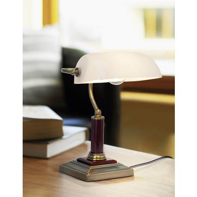 Table lamp with a white shade, illuminating a desk beside stacked books. A sofa and window are visible in the background.
