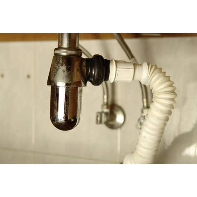 A chrome-plated siphon pipe beneath a sink, connected to a white, flexible waste pipe. Background: tiled wall.