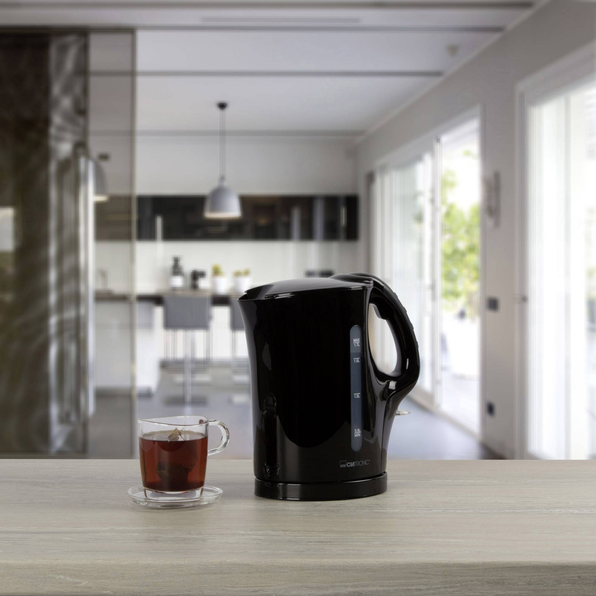 An electric kettle sits on a kitchen worktop beside a cup of tea in a modern kitchen with large windows in the background.