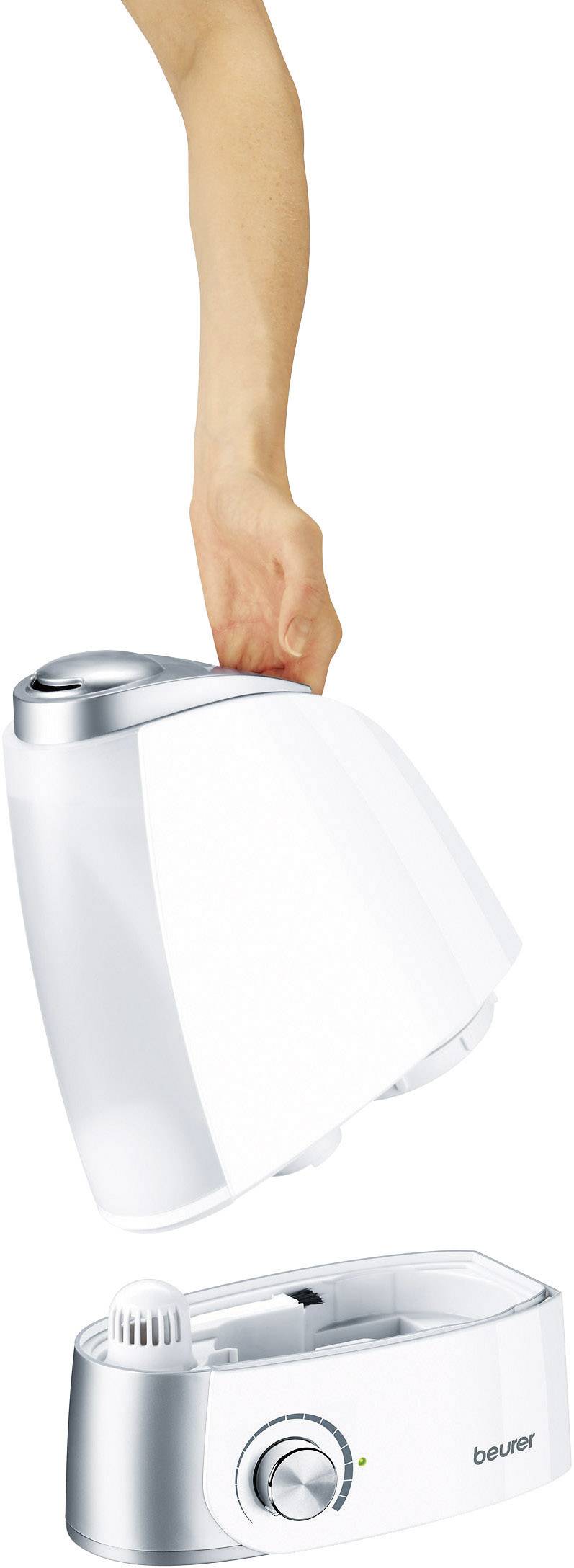 A hand lifts the water tank of a humidifier. The device has a silver-white design and a rotary control dial.