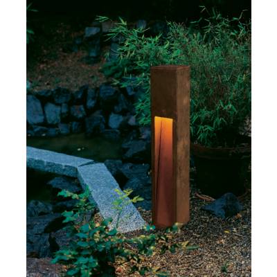 A wooden standard lamp stands in a garden, surrounded by green plants and stone pathways. It emits a warm light.