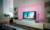 A modern living room with a flatscreen television in the centre. Bookshelves are positioned on the left side and photographs are displayed above. Warm ambient lighting creates a cosy atmosphere.