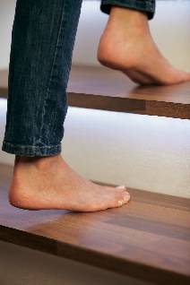 A person is walking barefoot up a wooden staircase. Their legs are dressed in blue jeans.