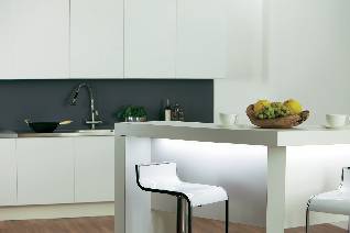 Modern kitchen with a white dining table and bar stools, fruit bowl and crockery. White wall cupboards and grey splashback behind the sink.