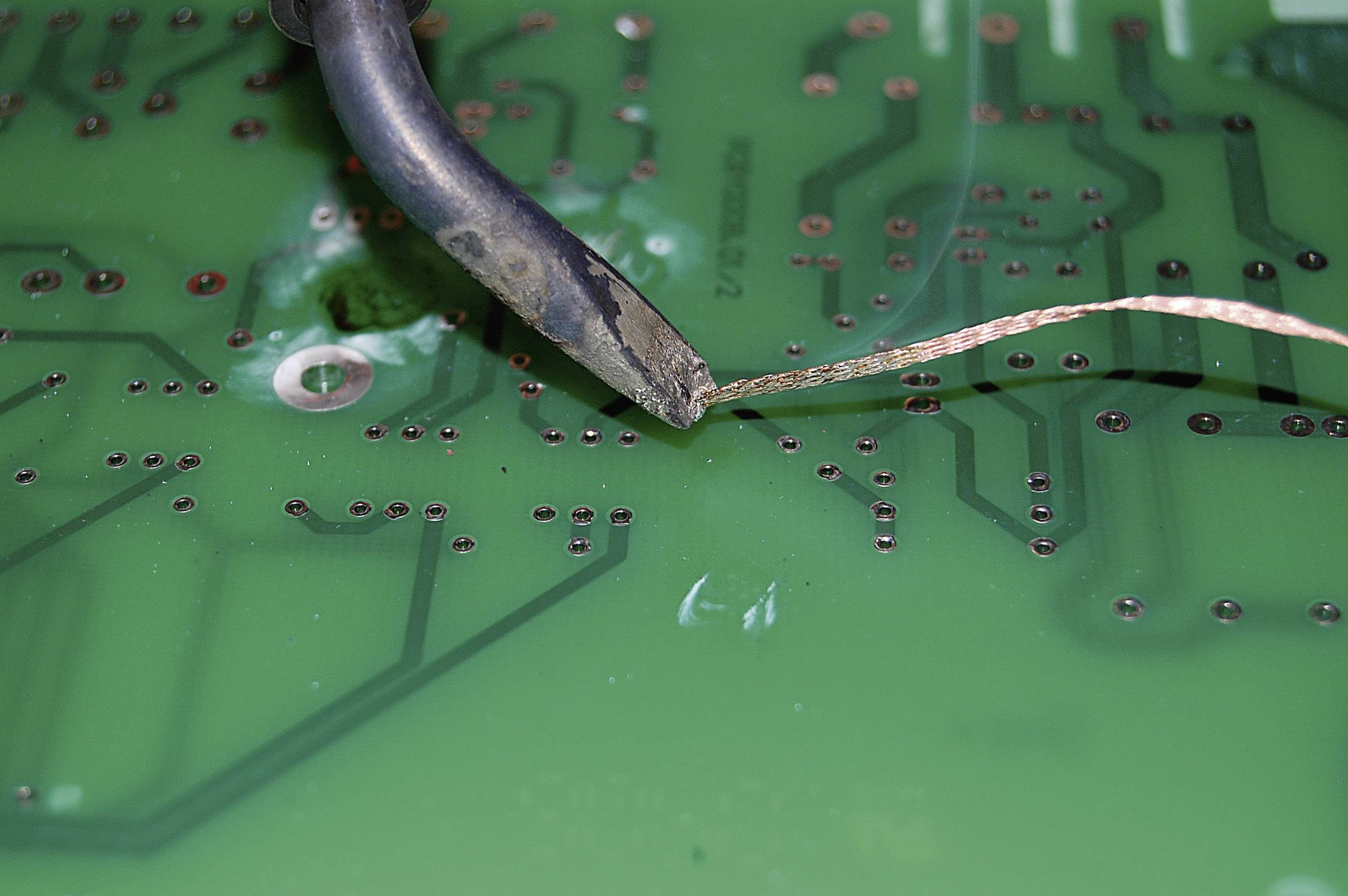 A soldering iron heats a circuit board to remove solder connections. Close-up shows circuit tracks and solder points.
