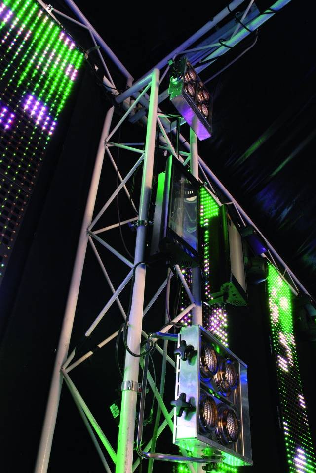 Stage lights and LED panels shine in green and purple, mounted on a frame at an event.
