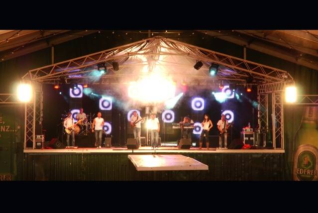 A band is playing on a lit stage with instruments against a background of circular light effects.