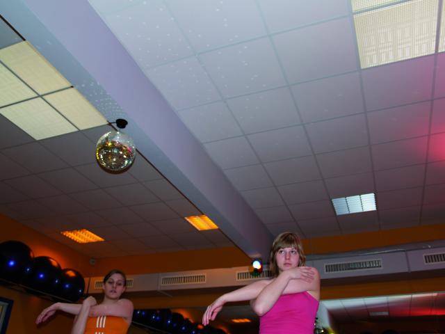 Two people in a gym performing dance moves, surrounded by mirrors, with a disco ball hanging from the ceiling.
