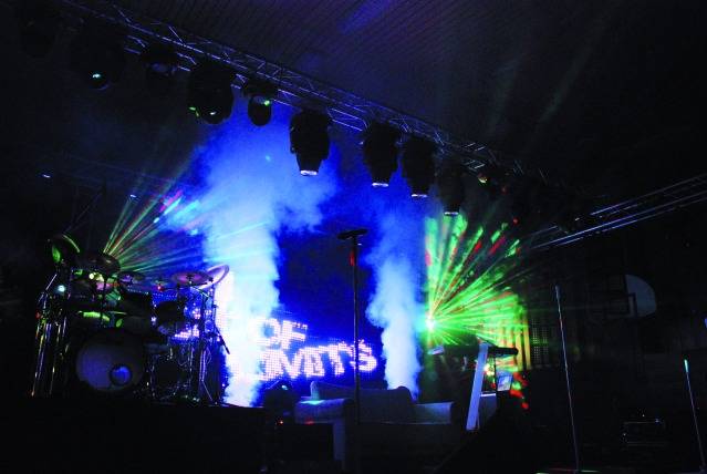 Stage with a drum kit, microphone stands, and colourful lights. Smoke and illumination create an energetic atmosphere.