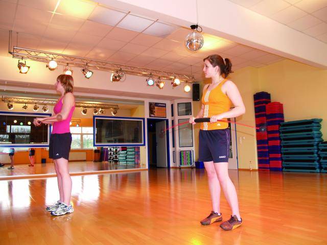 Two people are skipping with a rope in a gym. Mirrors in the background reflect the room, and colourful mats are stacked in piles.