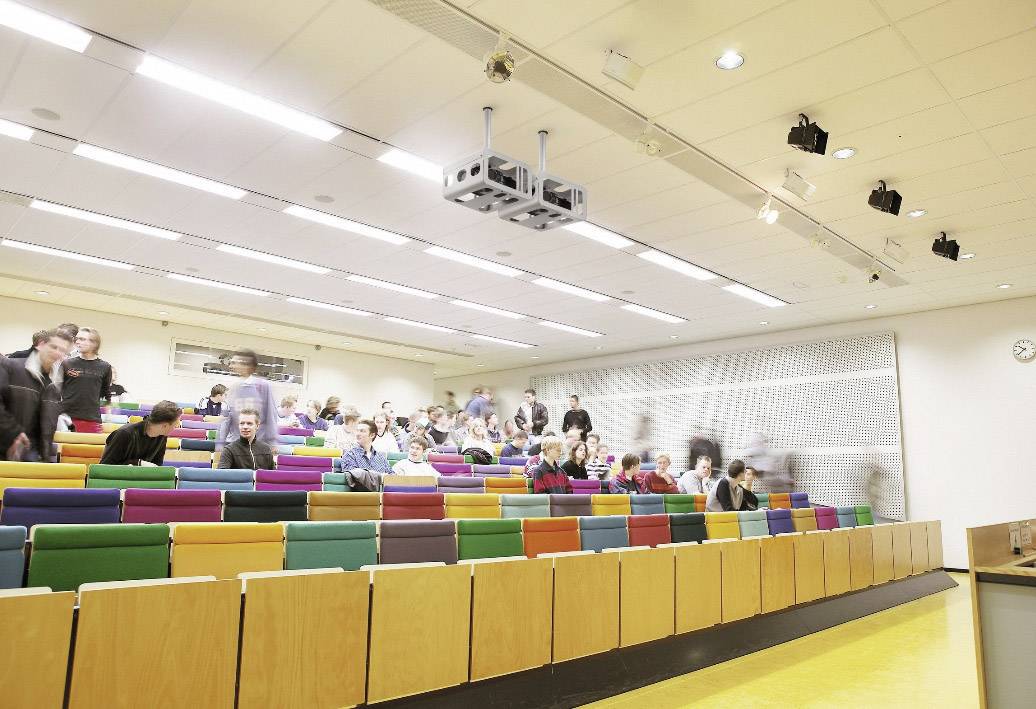 Empty lecture hall with colourful seats; some people are coming in or sitting down, with a clock on the wall in the background.