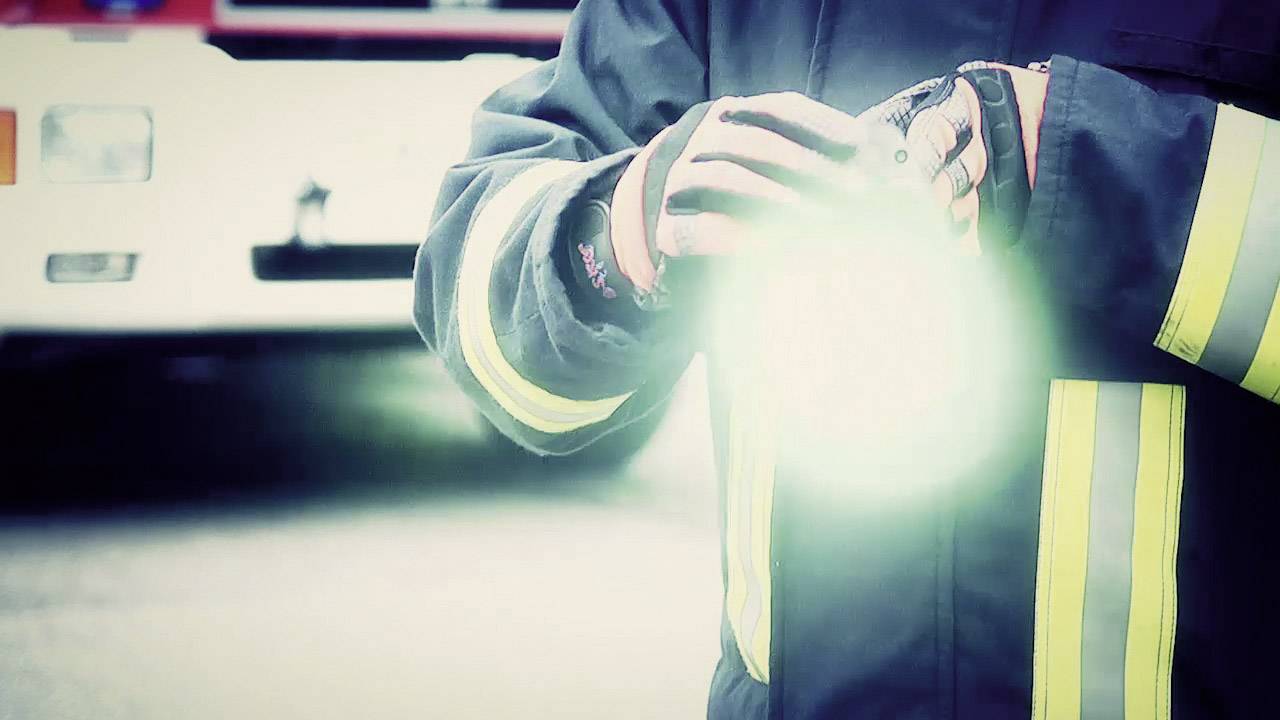 A firefighter in protective gear holds a bright torch, with a blurred fire engine in the background.