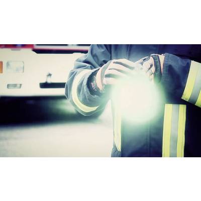 A firefighter in protective gear holds a bright torch, with a blurred fire engine in the background.