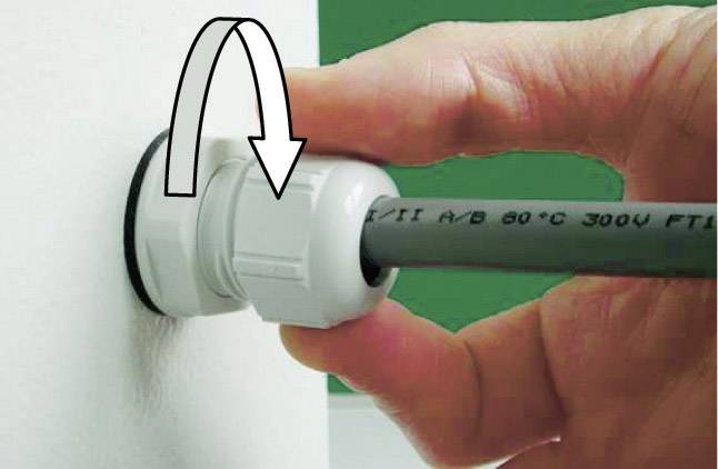 A hand pulls a cable through a white screw connection in the wall. An arrow indicates the direction of rotation for tightening. A plastic clamp secures the cable.