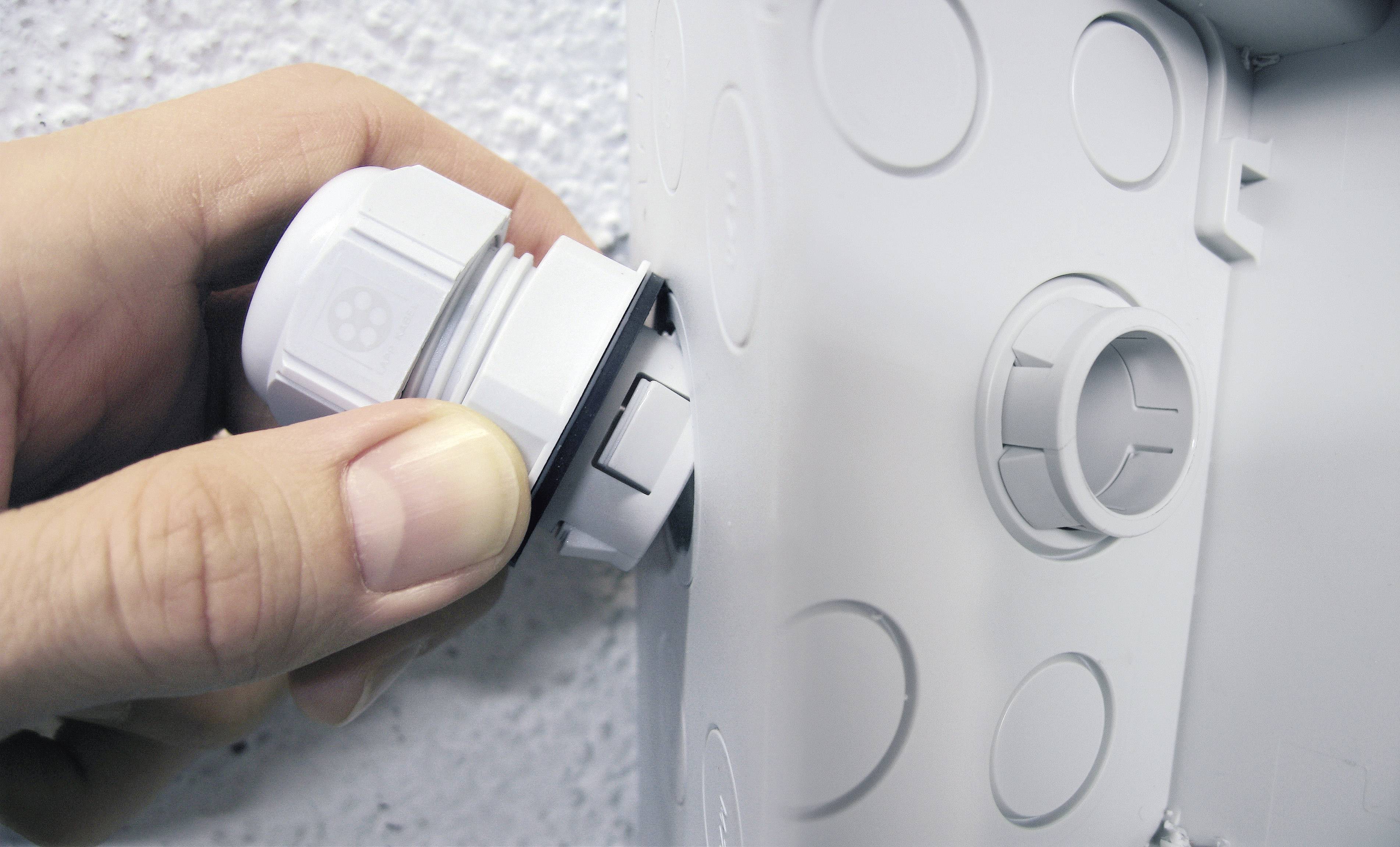 A hand is mounting a grey cable gland onto an enclosure that is fixed to a wall.