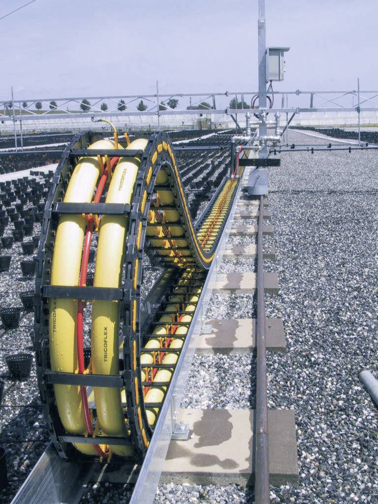 A cable drag chain on rails in an industrial setting, with empty planting containers on the left. The scene appears technical and orderly.