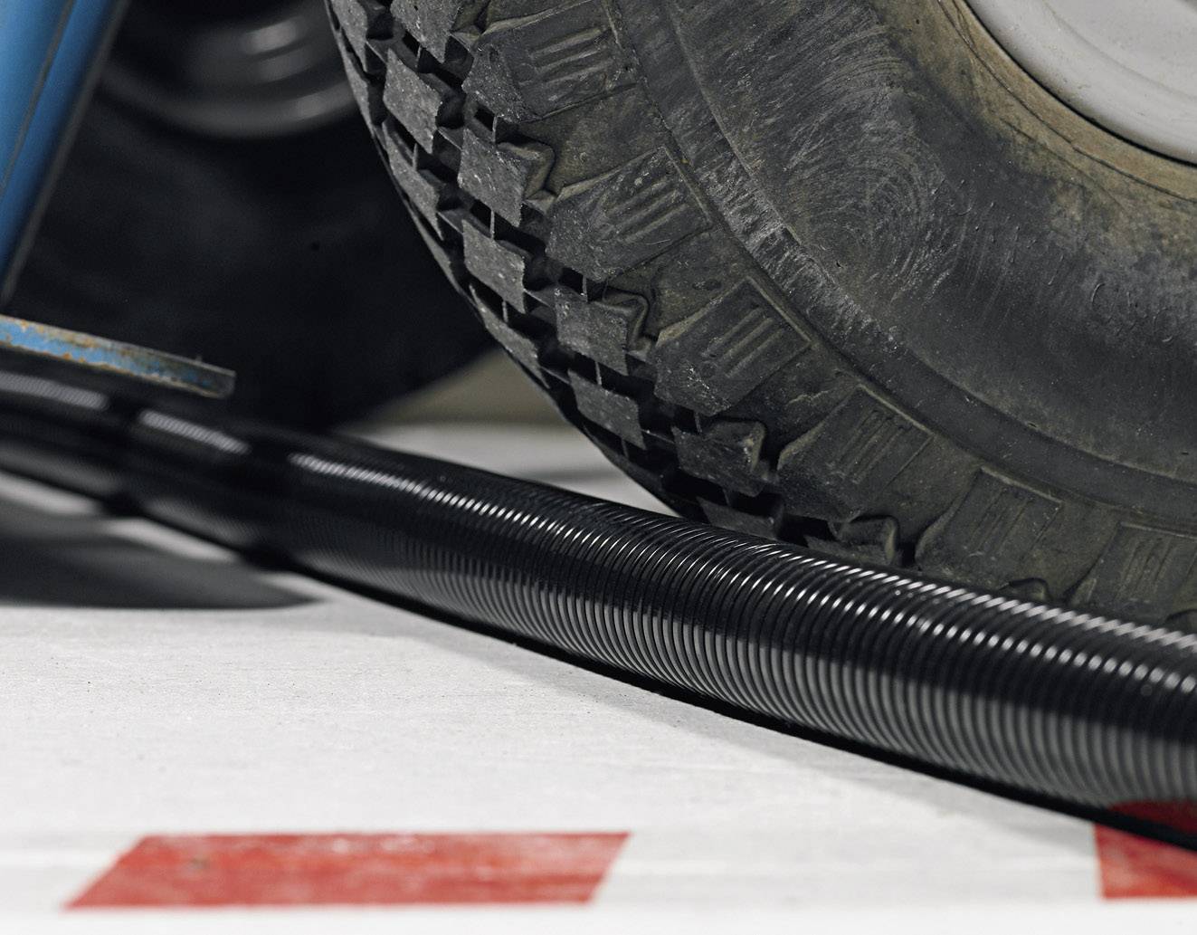 A heavy vehicle wheel runs over a black corrugated pipe on a red and white tiled floor.