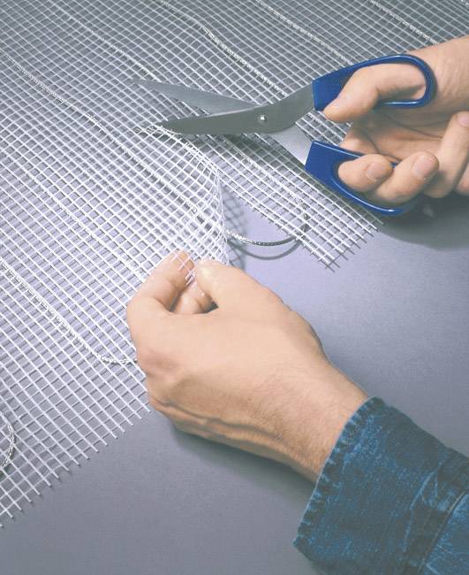 A person is cutting a mesh-like material with blue scissors. The hands are in focus and show the cutting process.