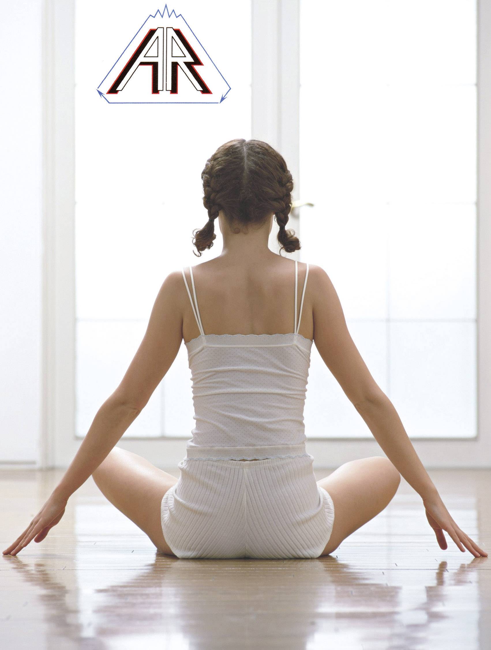 A woman is sitting cross-legged on a wooden floor in a bright room.